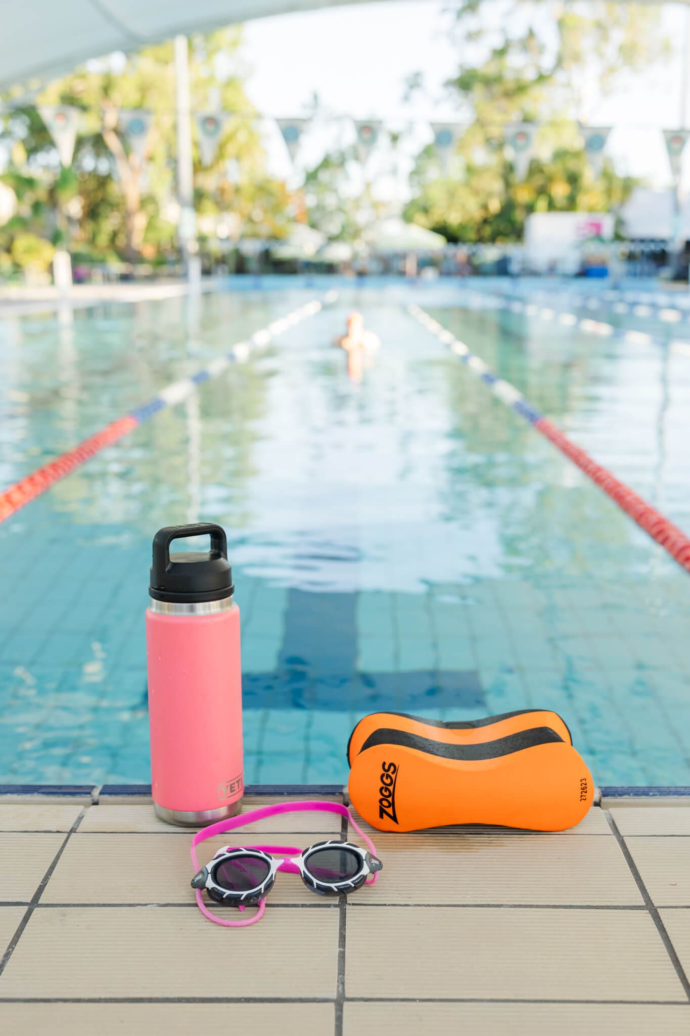 Bright and colourful image a water bottle, goggles and a floaty in the foreground, an Olympic sized pool in the background.