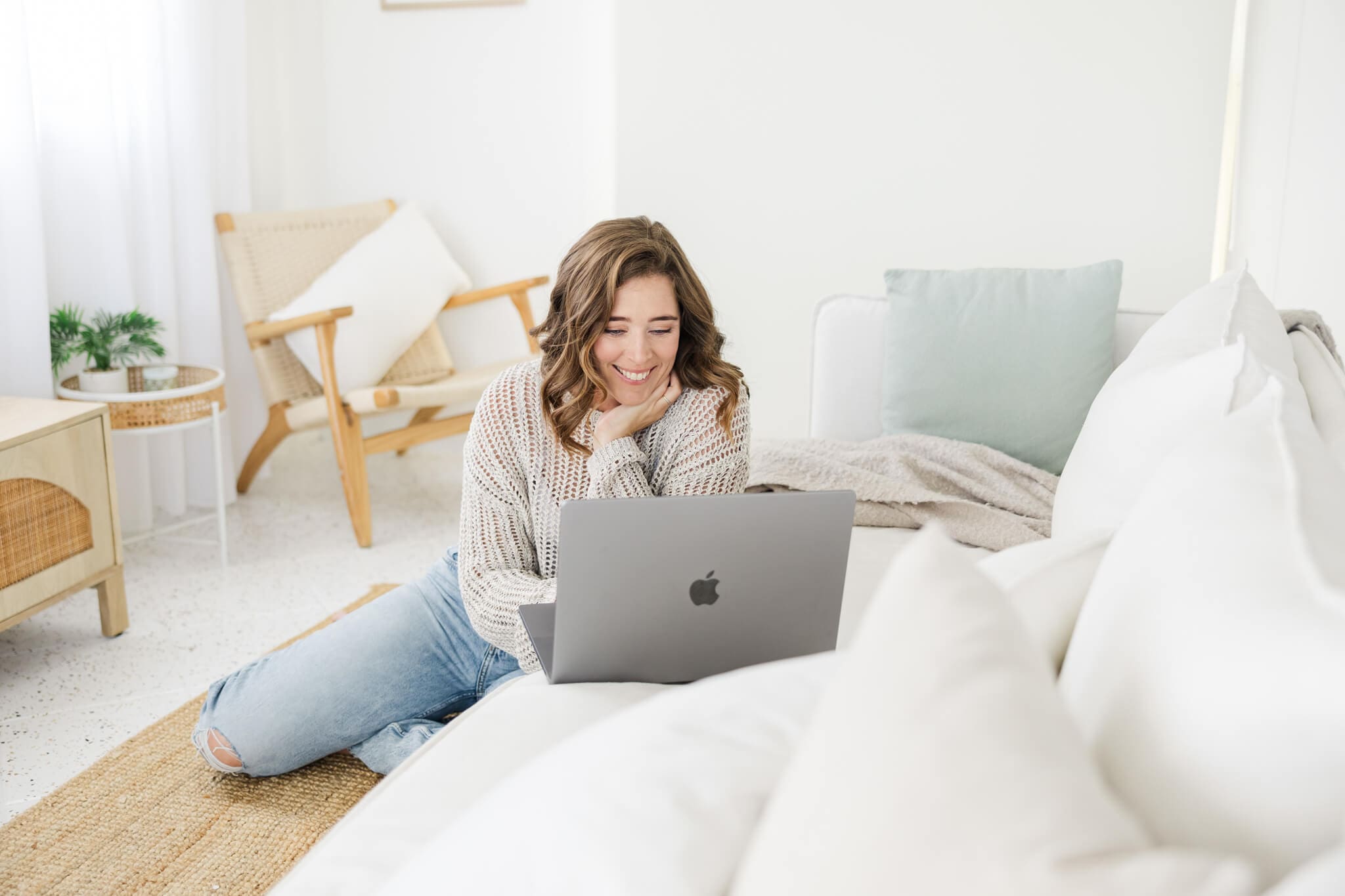 A woman sitting down on a lounge floor in a long sleeve top and blue jeans, looking at a laptop that is on the sofa.
