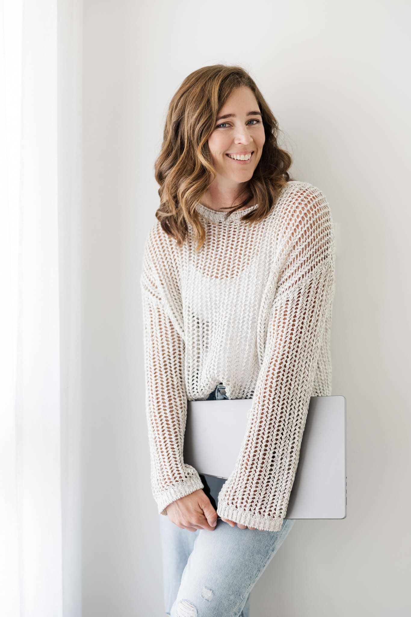 Woman leans against a wall, holding a laptop while she poses for her branding session, dressed in blue jeans.
