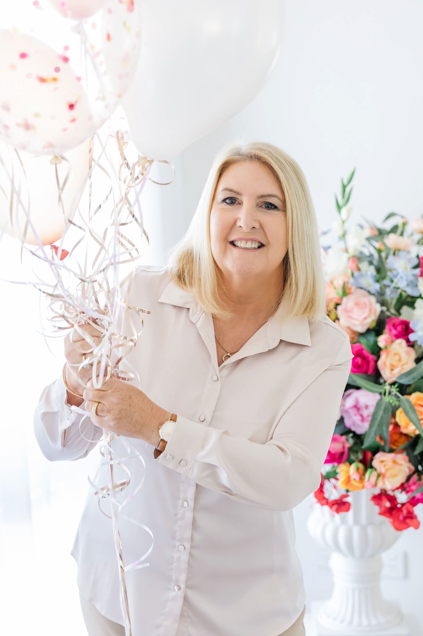 A woman in a white top and with blonde hair, poses with a bunch of balloons and a large bunch of flowers in the background.