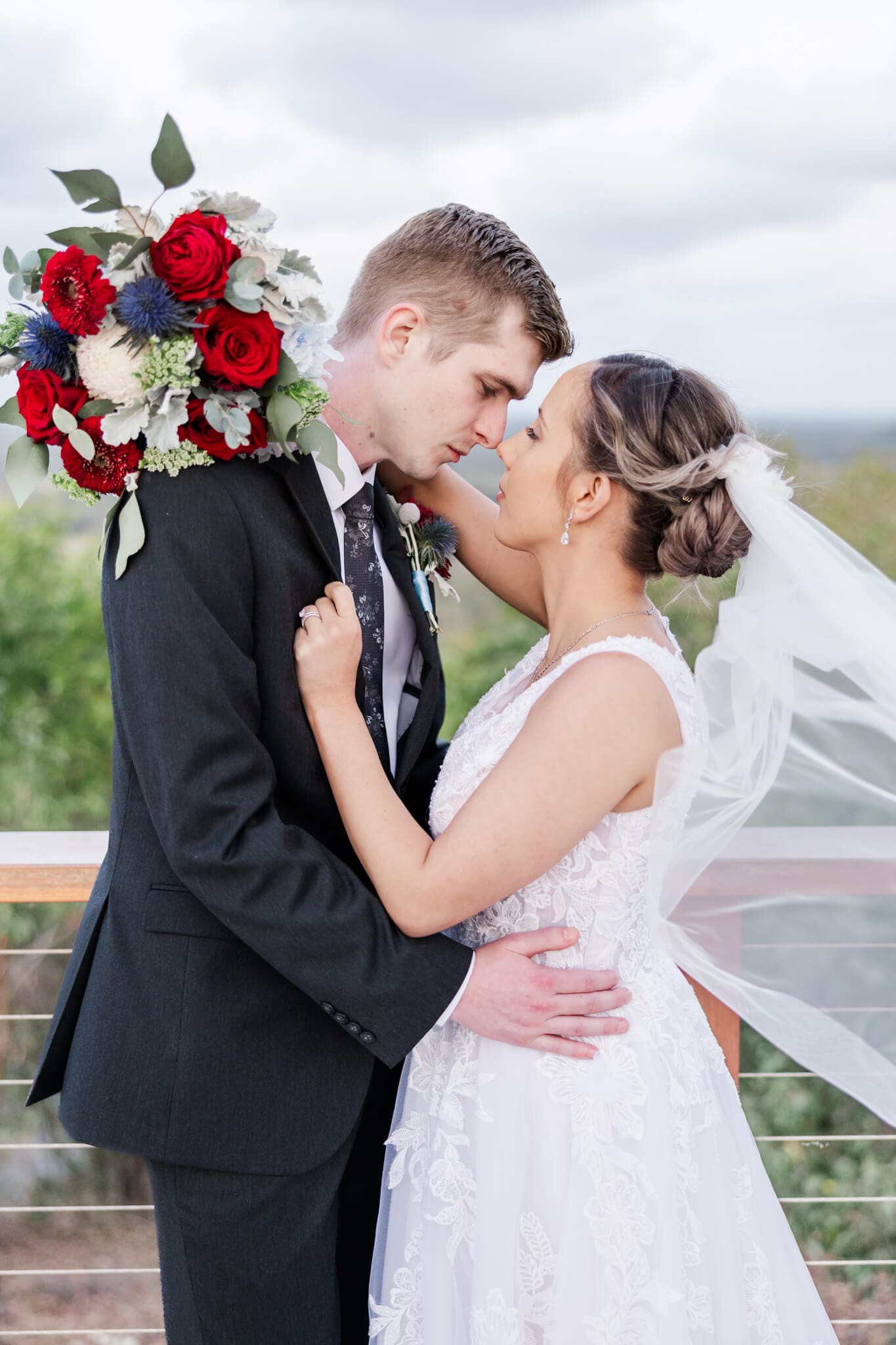 A bride and groom in each others arms, against a wooden railing while the bride's veil flys in the wind and she holds a stunning bouquet created by Yeppoon florist Dusty Blooms.