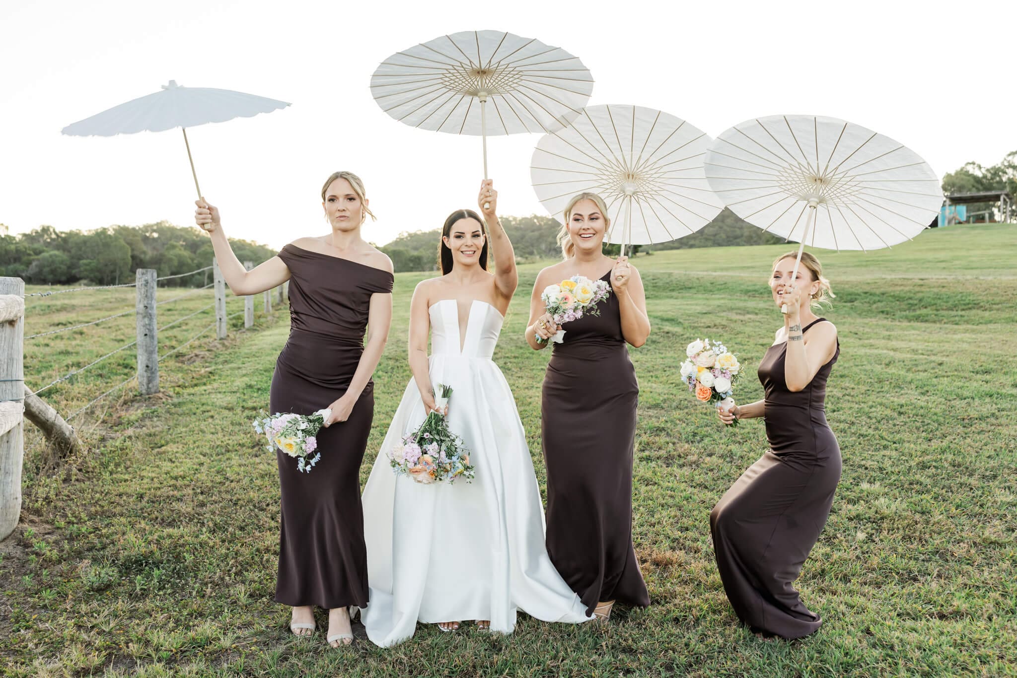 A bride and her bridesmaids dance with paper umbrellas in an open paddock.