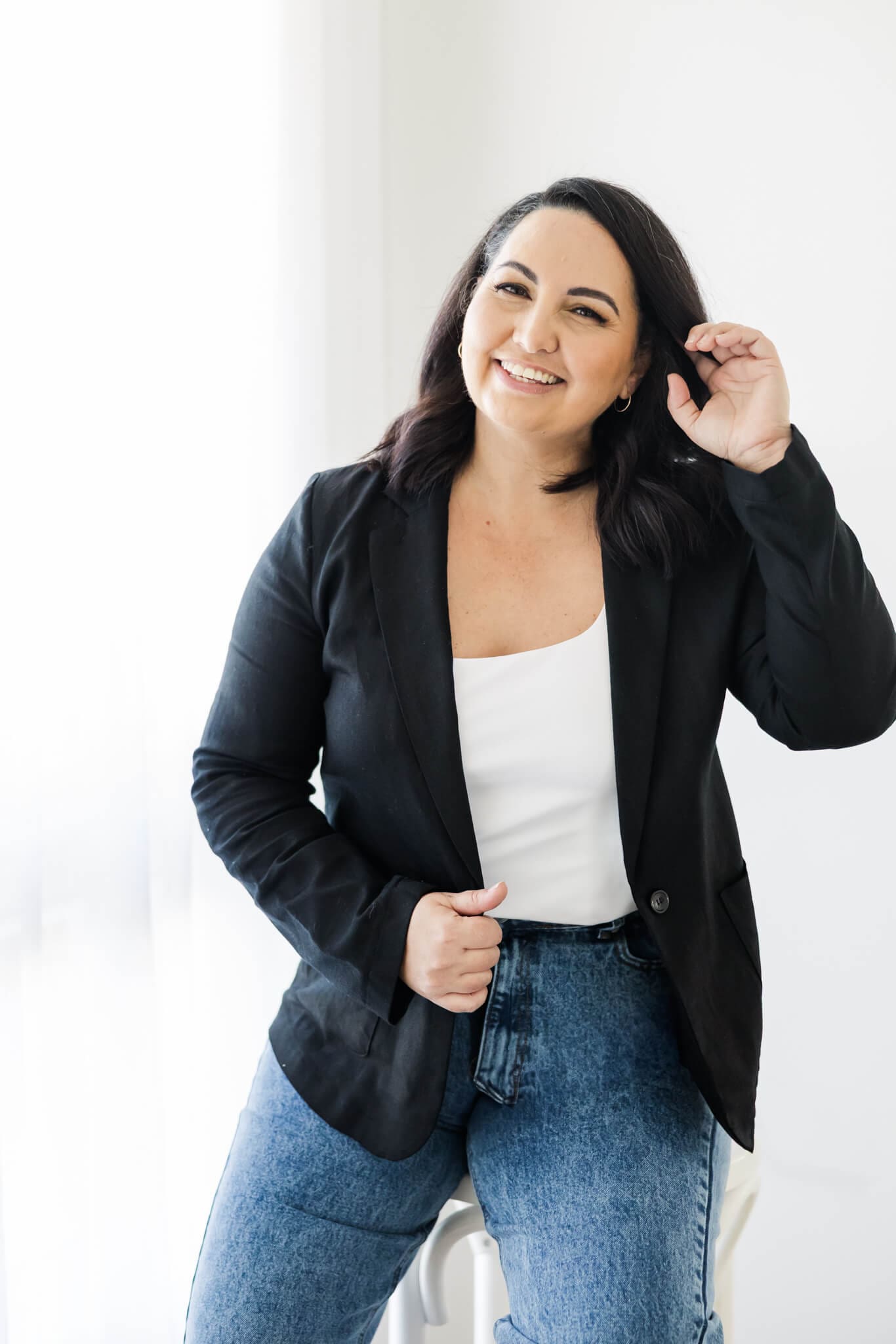 Branding photo of an event planner, seated at a stool, brushing her hair behind her ears, wearing blue jeans and a white top overlaid with a black jacket.
