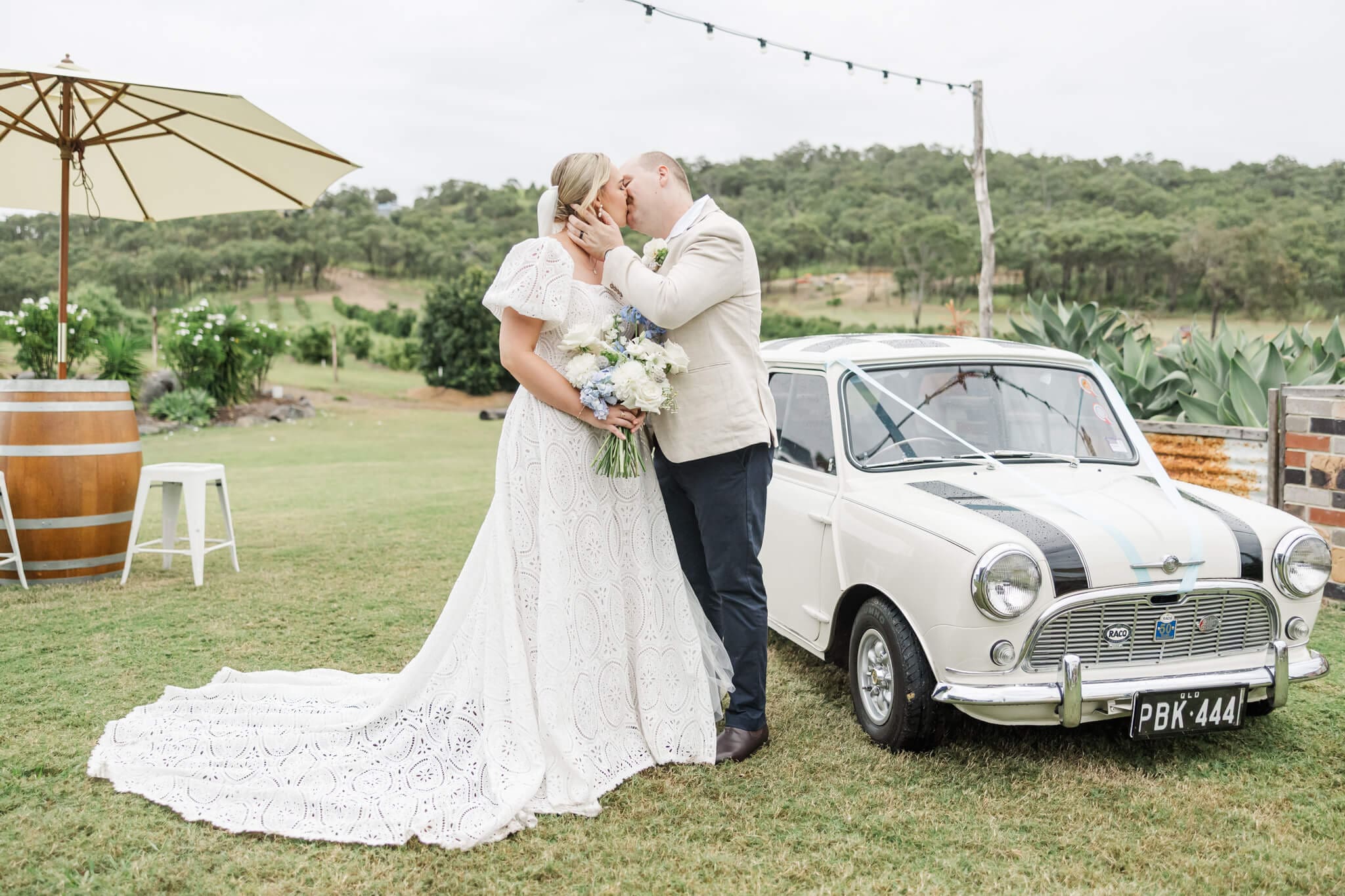 Bride and groom kiss near an old fashioned car while the bride holds her Posy Yeppoon bouquet.
