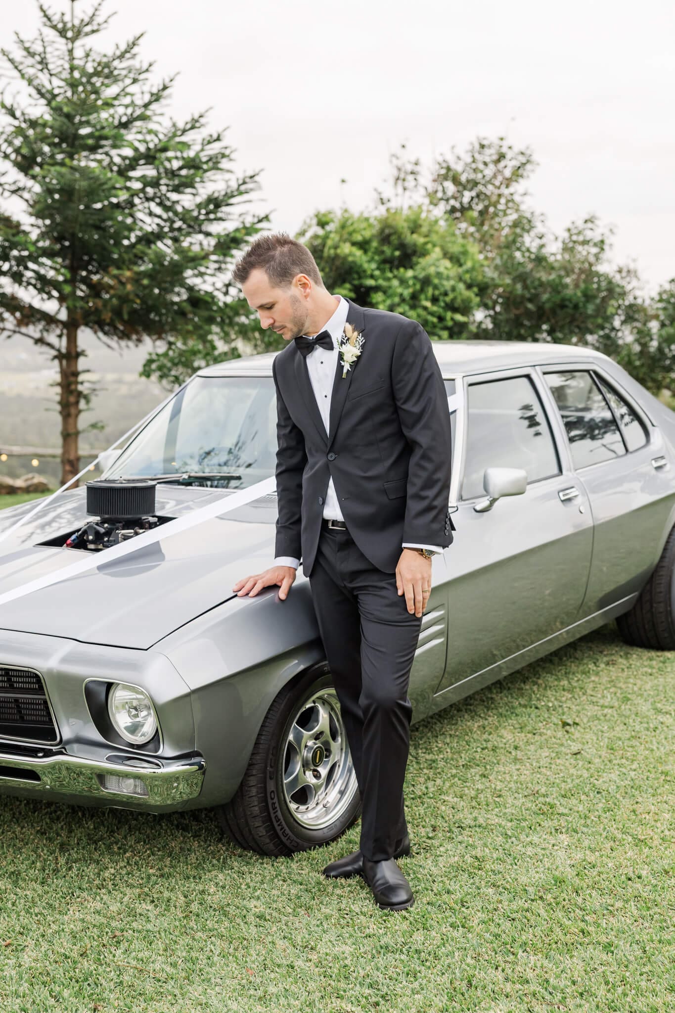 Groom glancing down at a vintage car, showcasing a mens suit from Rockhampton.