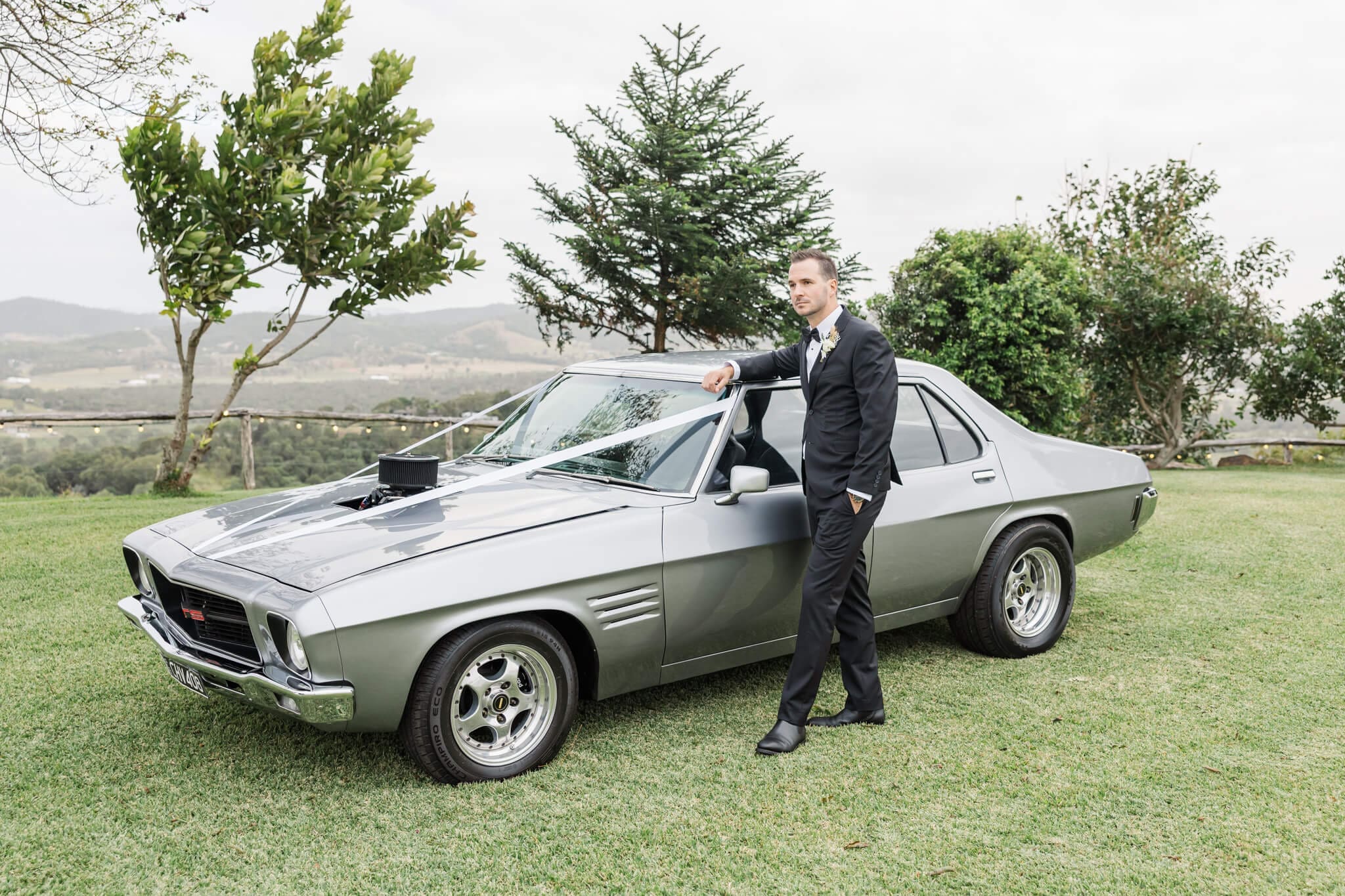Groom stands with a vintage car while dressed in a suit from Rockhampton menswear stores.