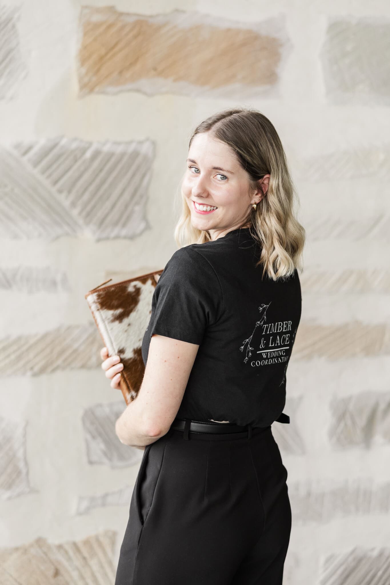 Timber and Lace Coordination owner, Shelby, smiles back over her shoulder at the camera, while holding her wedding planner folder.