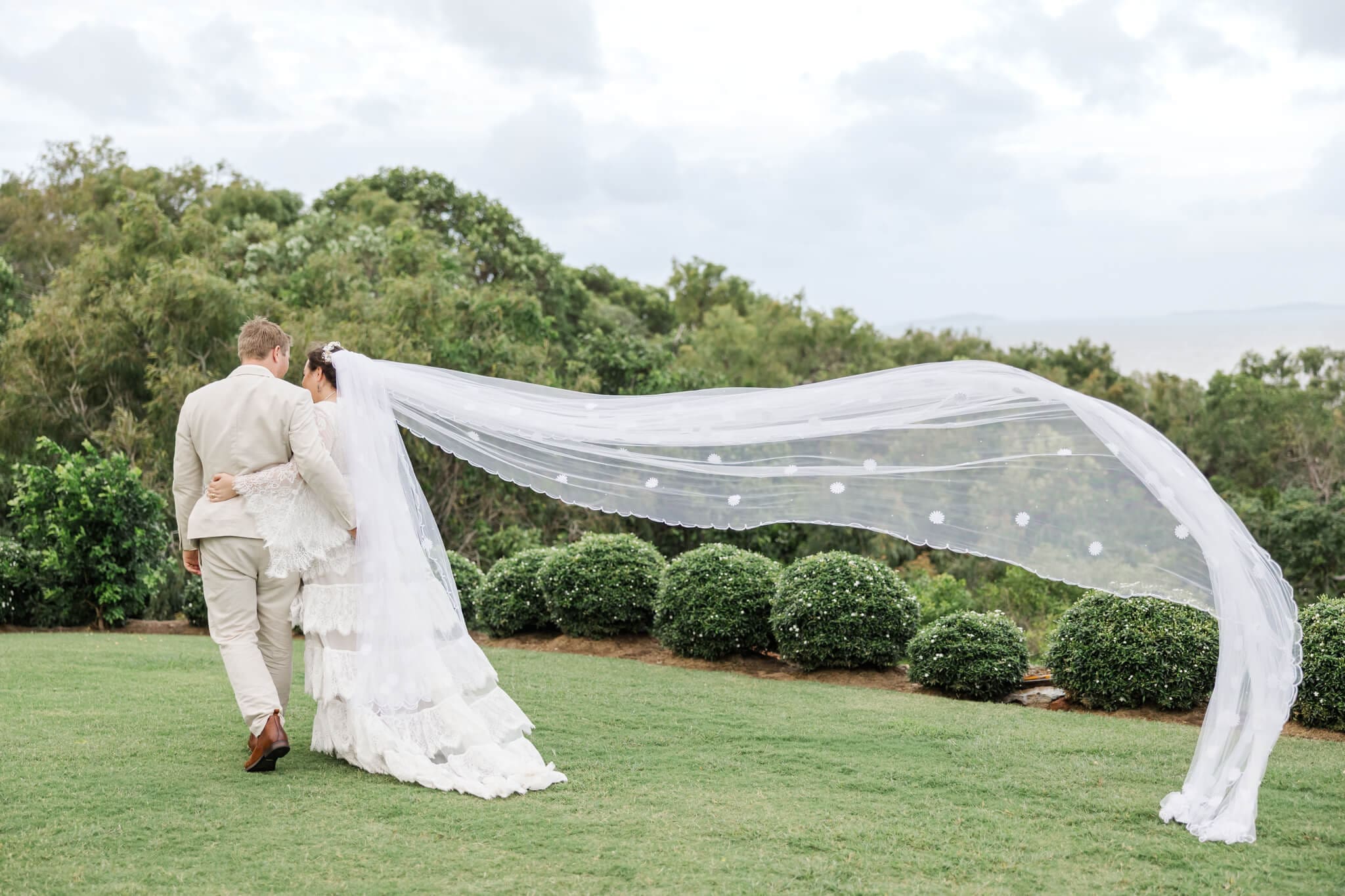 Bride and groom walk away from the camera with the extra long veil flowing back in the wind.